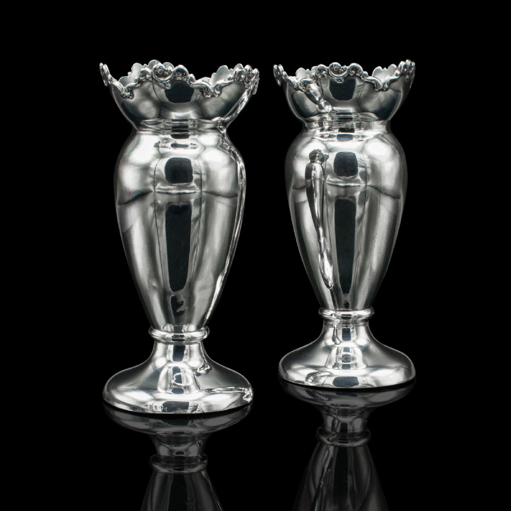 A pair of Edwardian hallmarked silver duck egg cups featuring crimped rims and pedestal bases.
