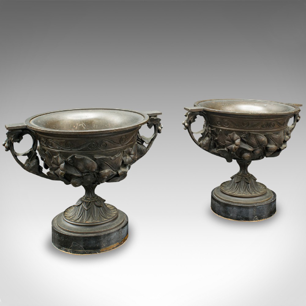 A pair of antique cast bronze goblets with high floral relief moulding resting on circular dark bases.