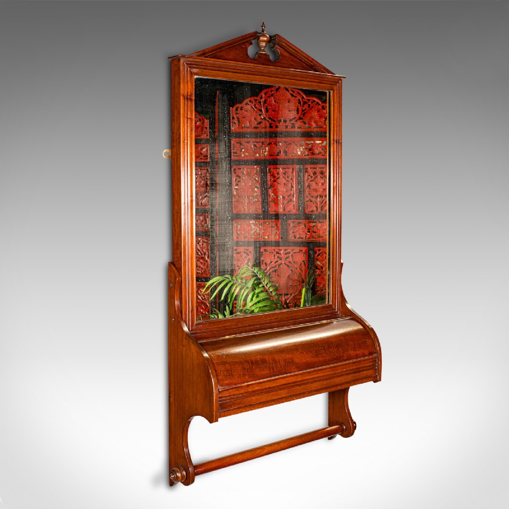 A Victorian mahogany wall-mounted valet mirror featuring a pediment top, rectangular glass plate, cylinder glove box, and lower hanging rail.