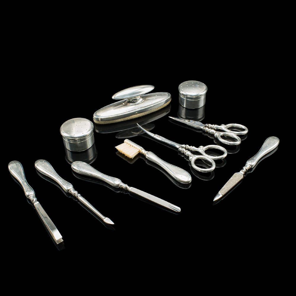 An eleven-piece Edwardian silver travelling grooming set arranged against a dark reflective surface, including scissors, nail files, brushes, and small lidded pots with punchwork detailing.