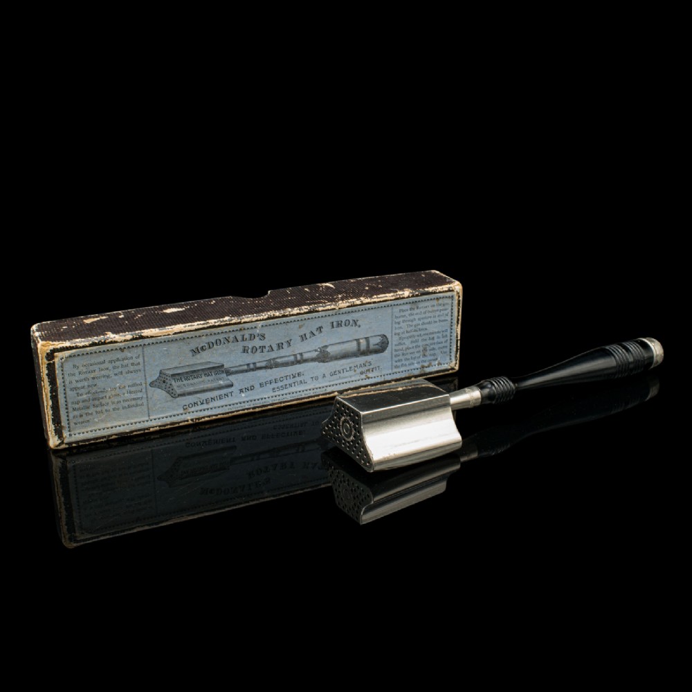 A nickel-plated McDonalds rotary hat iron with a black turned handle displayed next to its original printed cardboard box against a dark reflective surface.