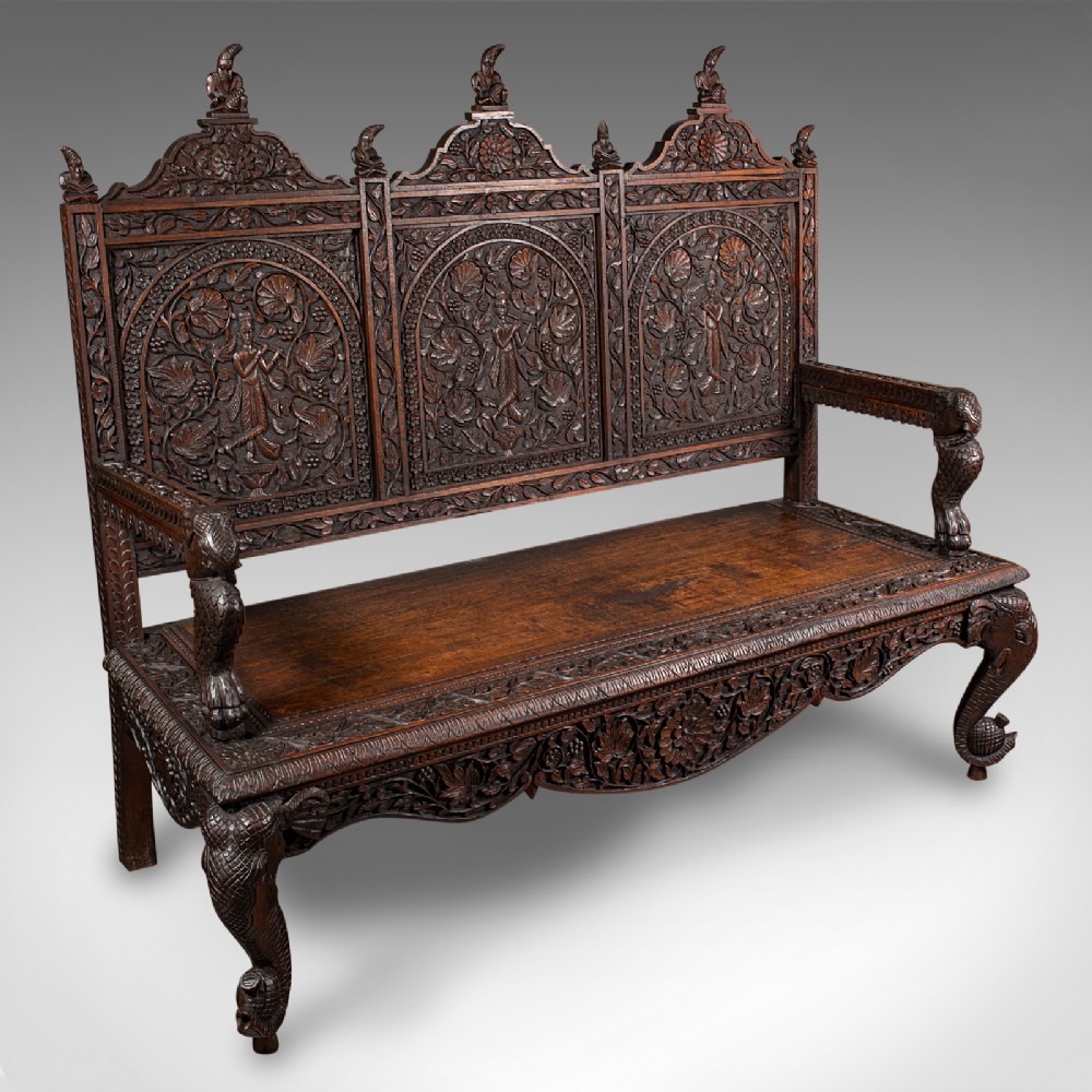 A large dark teak three-seat bench featuring arched back panels with relief carvings, beast-form armrests, and carved legs.