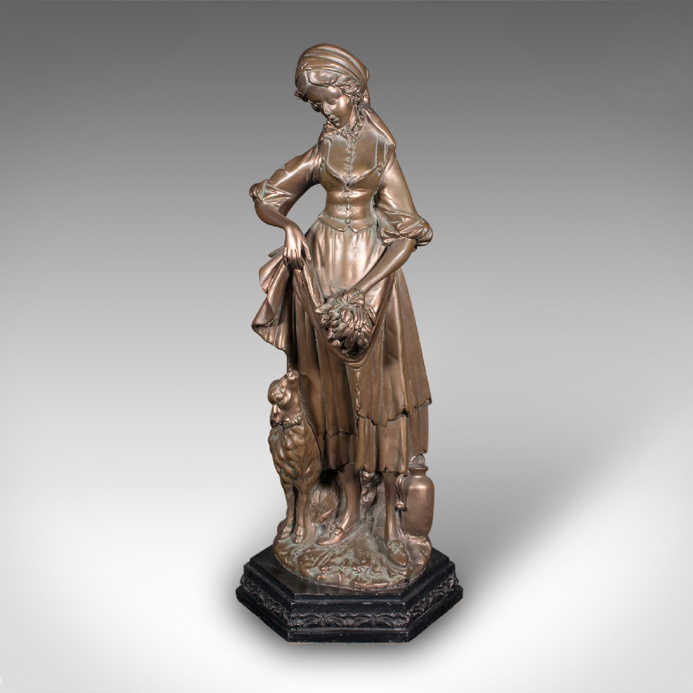 An antique bronzed plaster statue depicting a standing woman in rustic attire holding a basket alongside a small dog on a dark octagonal plinth.