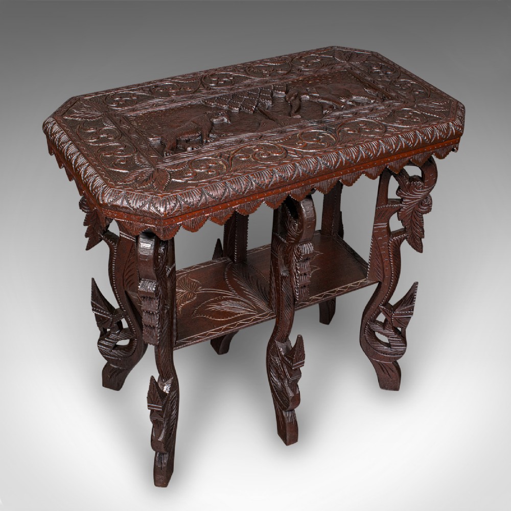 A dark brown hand-carved teak occasional table featuring animal relief carving on top, pierced foliage legs, and a lower shelf tier.