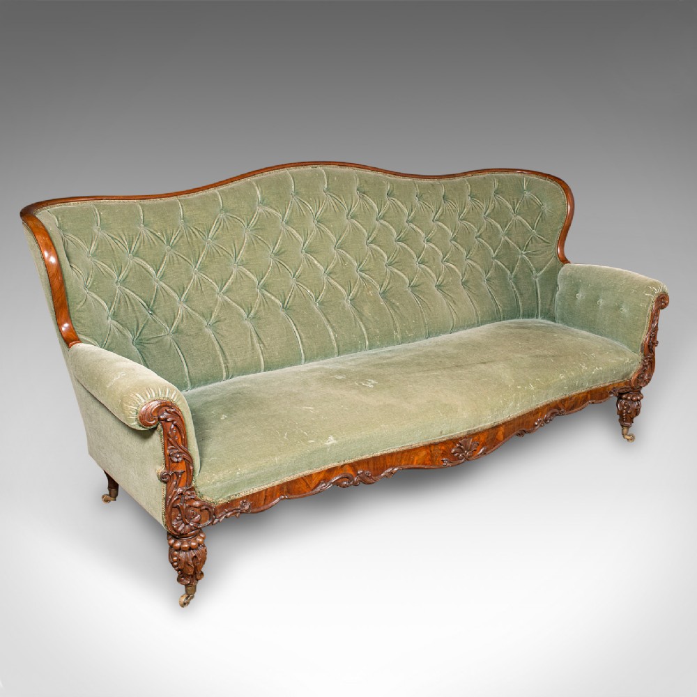 An antique seven-foot wide English settee featuring sage green velvet button-back upholstery, a carved walnut frame, and scrolled legs on brass castors.