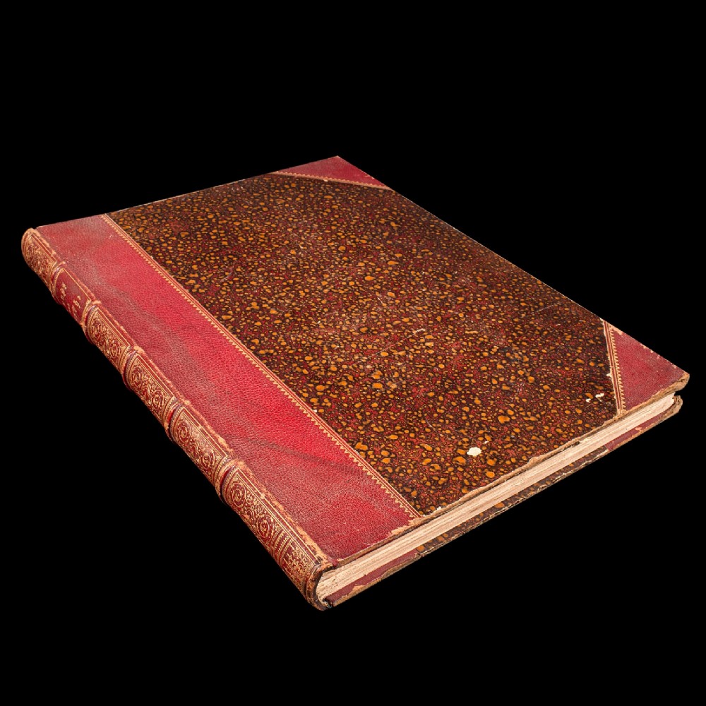 A large antique book bound in red leather and marbled brown paper with gold gilt detailing along the spine.