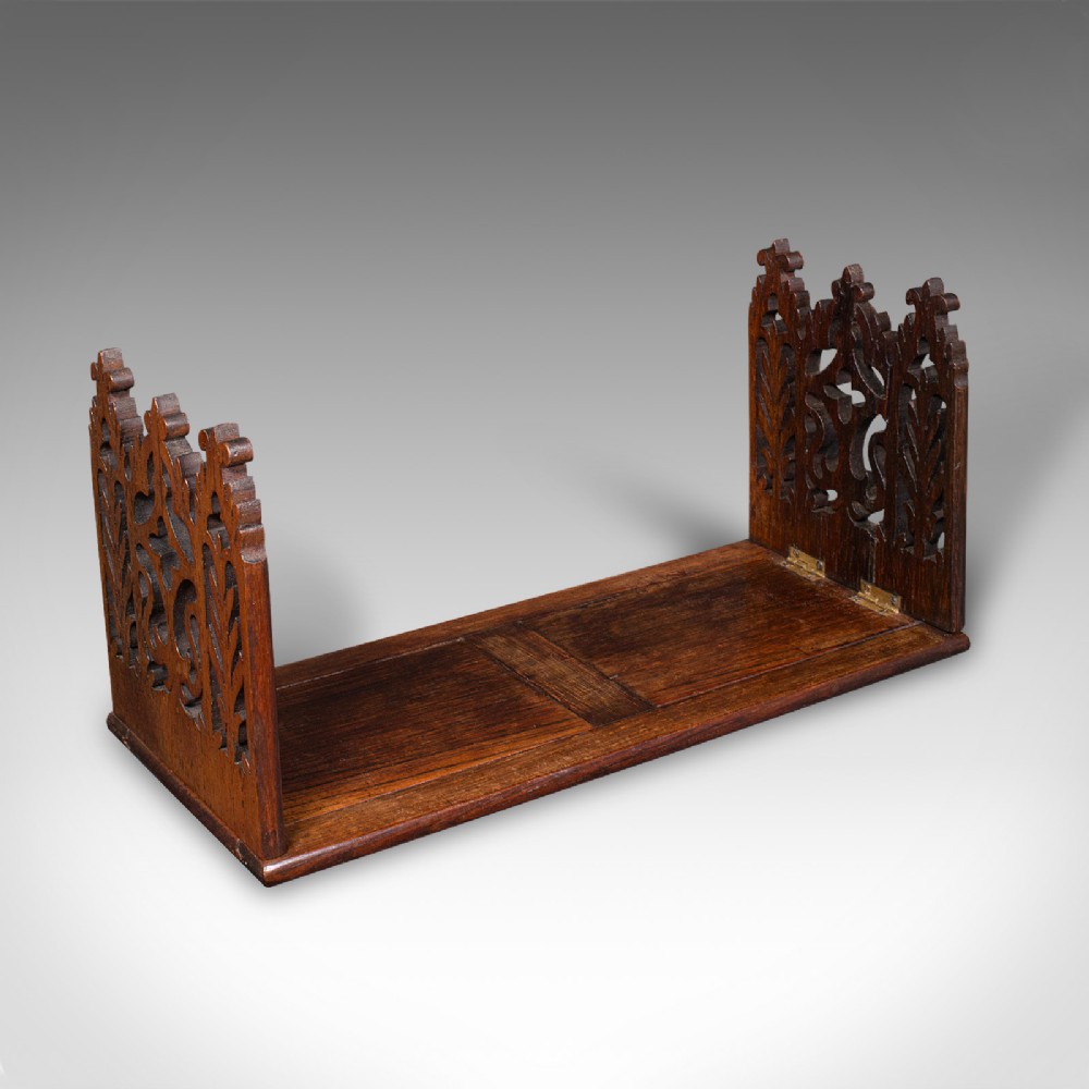 A dark oak Victorian extending book slide featuring pierced Gothic fretwork carved ends hinged to an adjustable base.