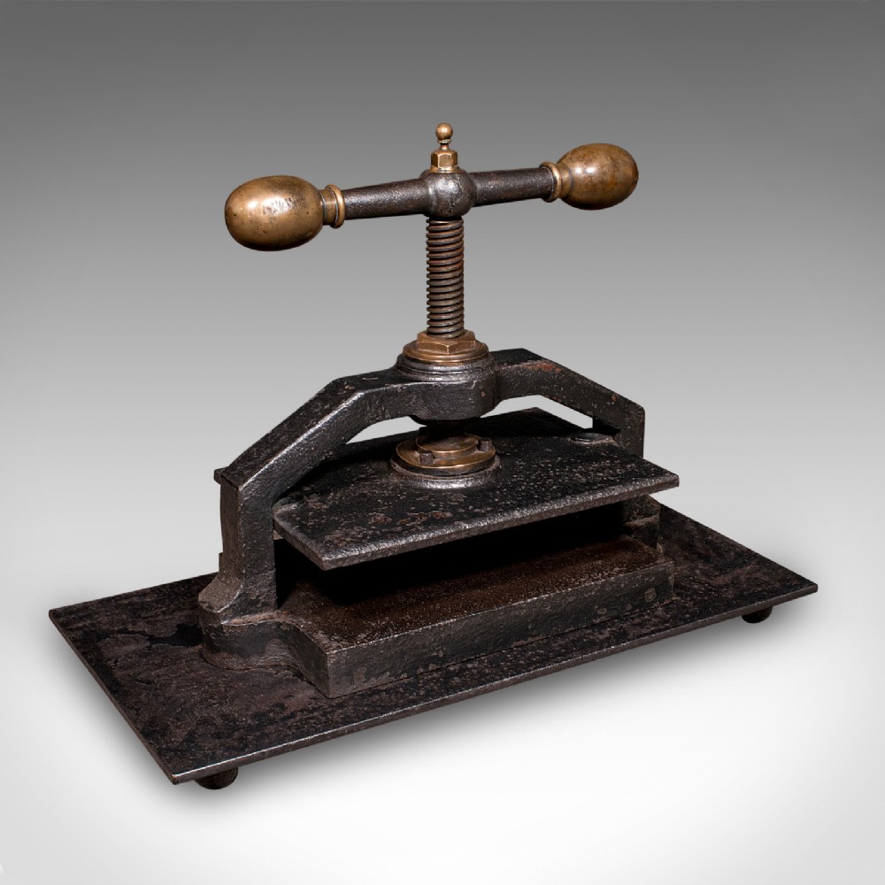 A dark cast iron book press featuring a central threaded screw, brass handle knobs, and a rectangular base plate.