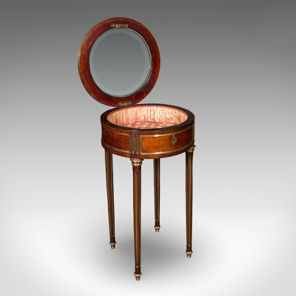An antique French circular bijouterie table crafted from kingwood, featuring fluted legs with brass mounts and an open hinged glazed lid revealing a tufted pink silk interior.