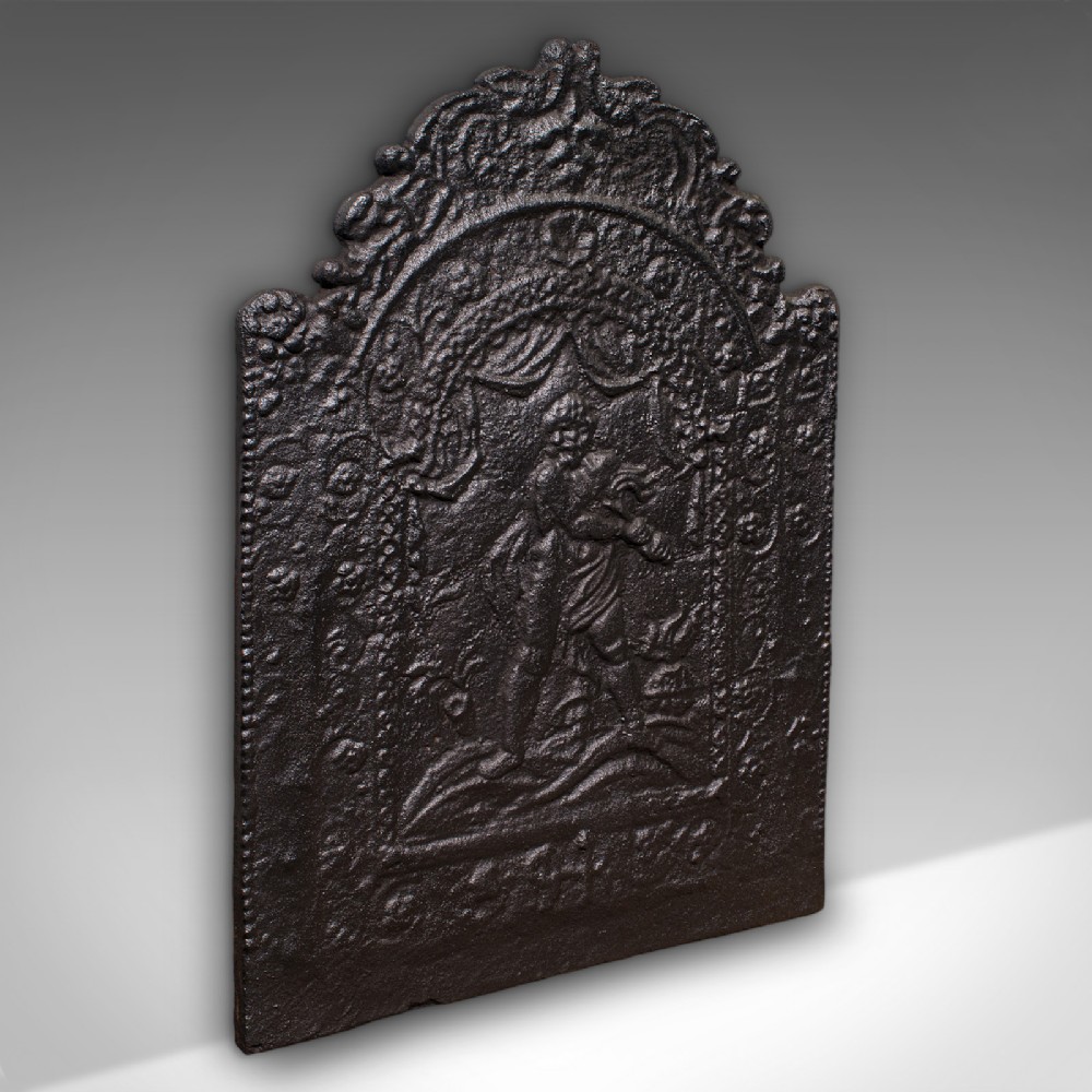 An arched dark cast iron fireback with relief carving of a classical figure framed by scrollwork.
