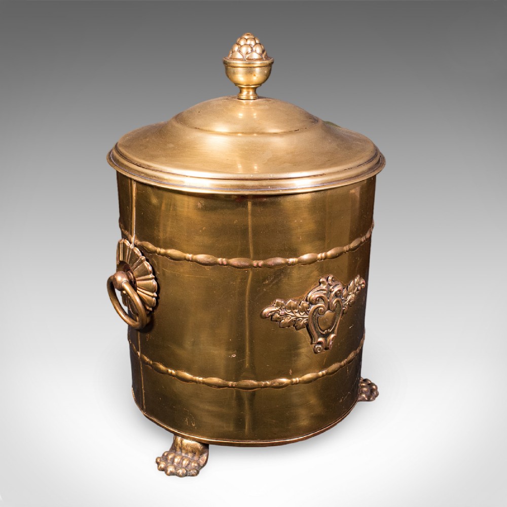 A cylindrical brass coal bin featuring a domed lid with a finial, side ring handles, embossed scrollwork, and lion paw feet.