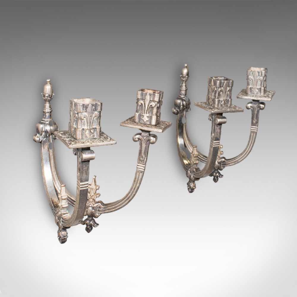 A pair of antique French silvered metal twin-branch candle sconces with fleur-de-lis detailing and square sockets.