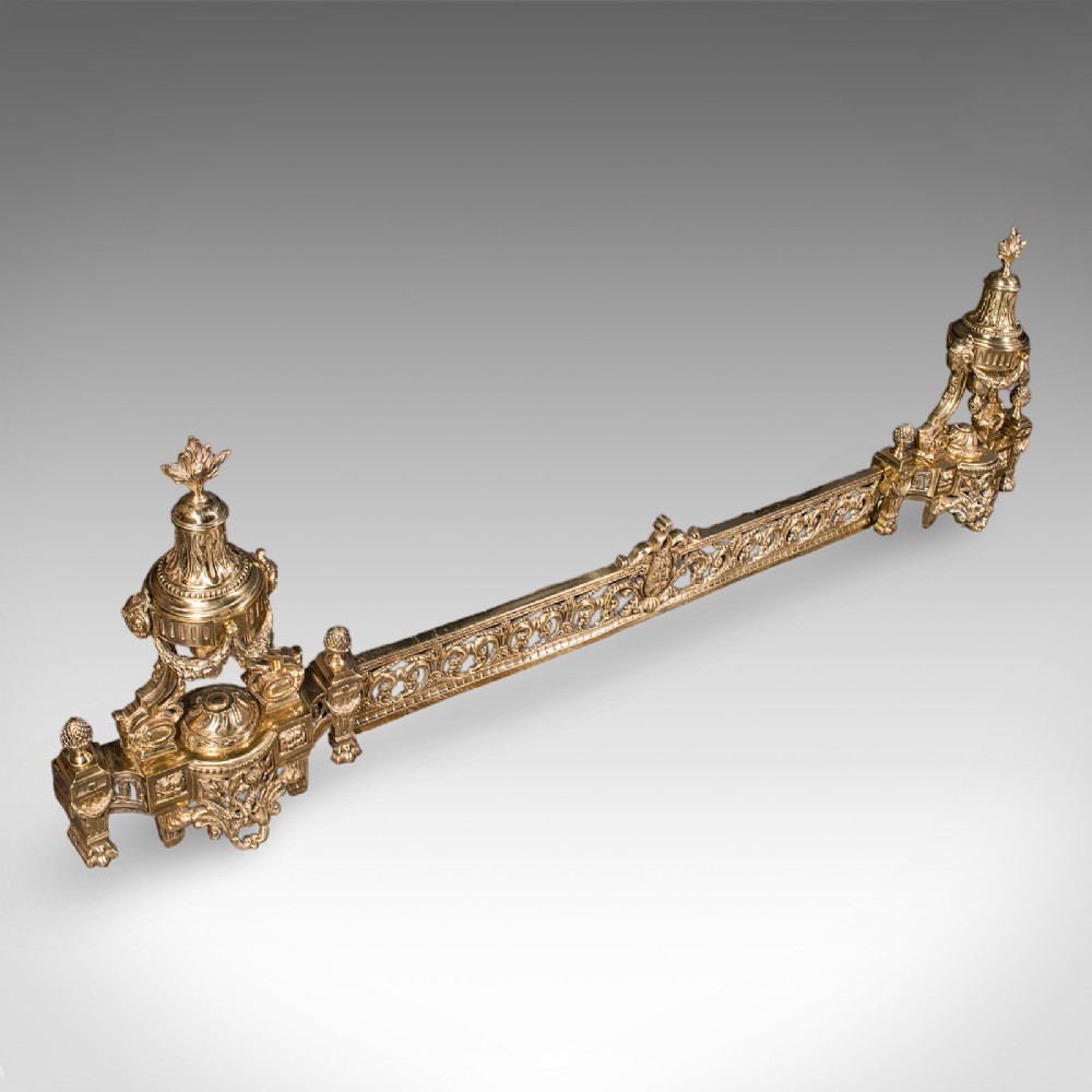 An ornate Victorian adjustable brass hearth fender featuring pierced scrollwork and classical urn finials on each end.