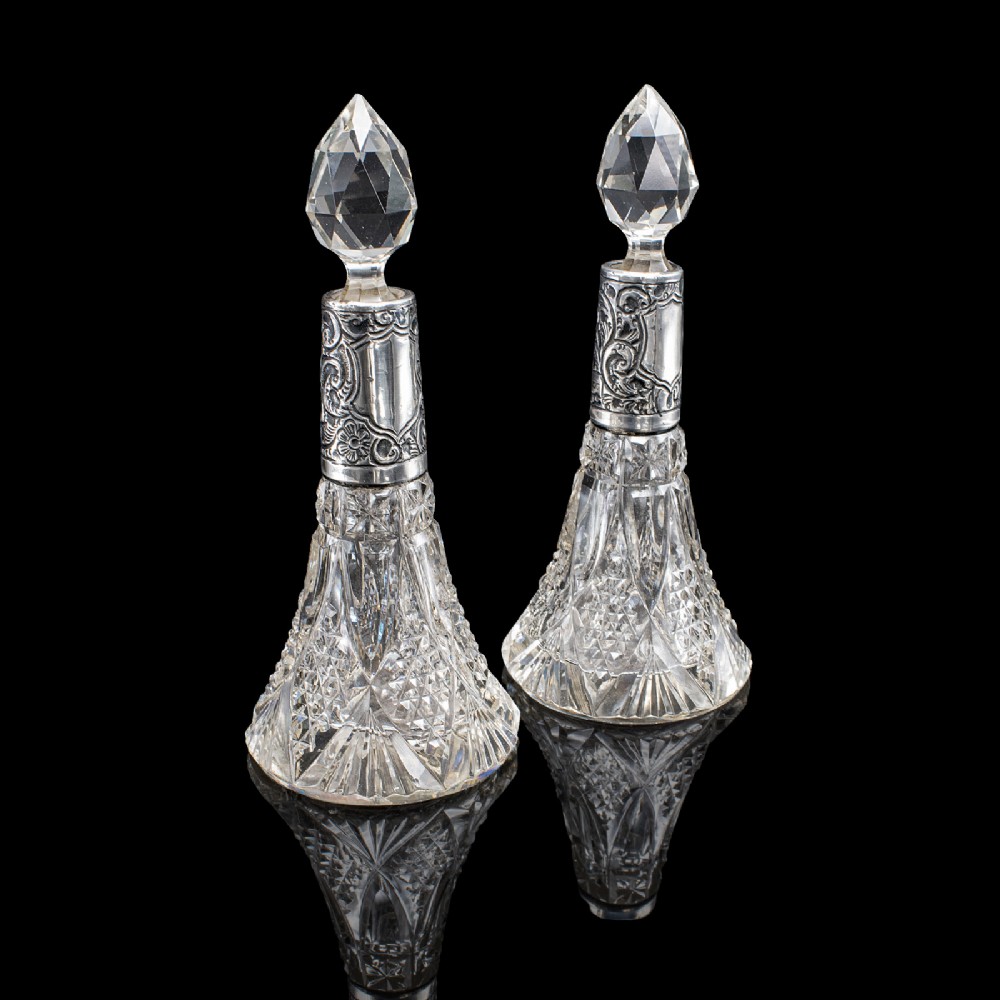 A pair of conical cut crystal scent bottles featuring embossed silver collars and faceted glass stoppers.