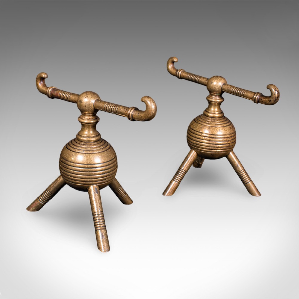A pair of late Victorian brass fireside tool rests featuring ribbed spherical central bodies, tripod legs, and hooked horizontal crossbars.