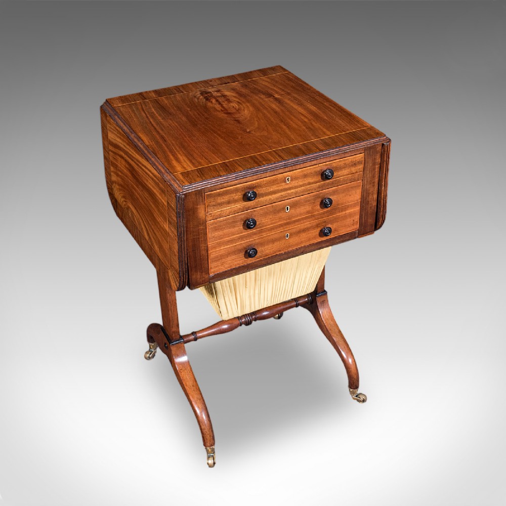 A Regency mahogany drop-leaf sewing table with three drawers, a pleated cream fabric work basket, and swept legs on brass castors.