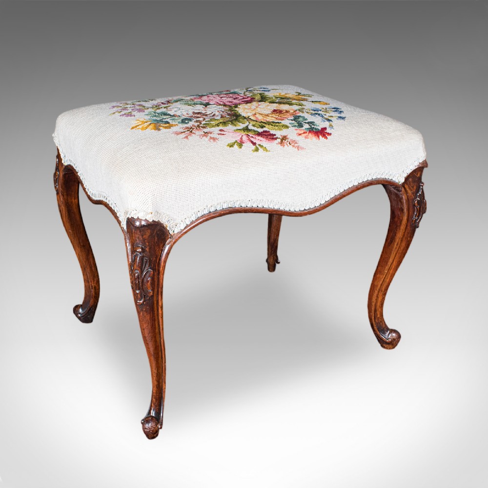 An Early Victorian walnut footstool featuring cabriole legs and a cream upholstered top detailed with a floral needlepoint design.