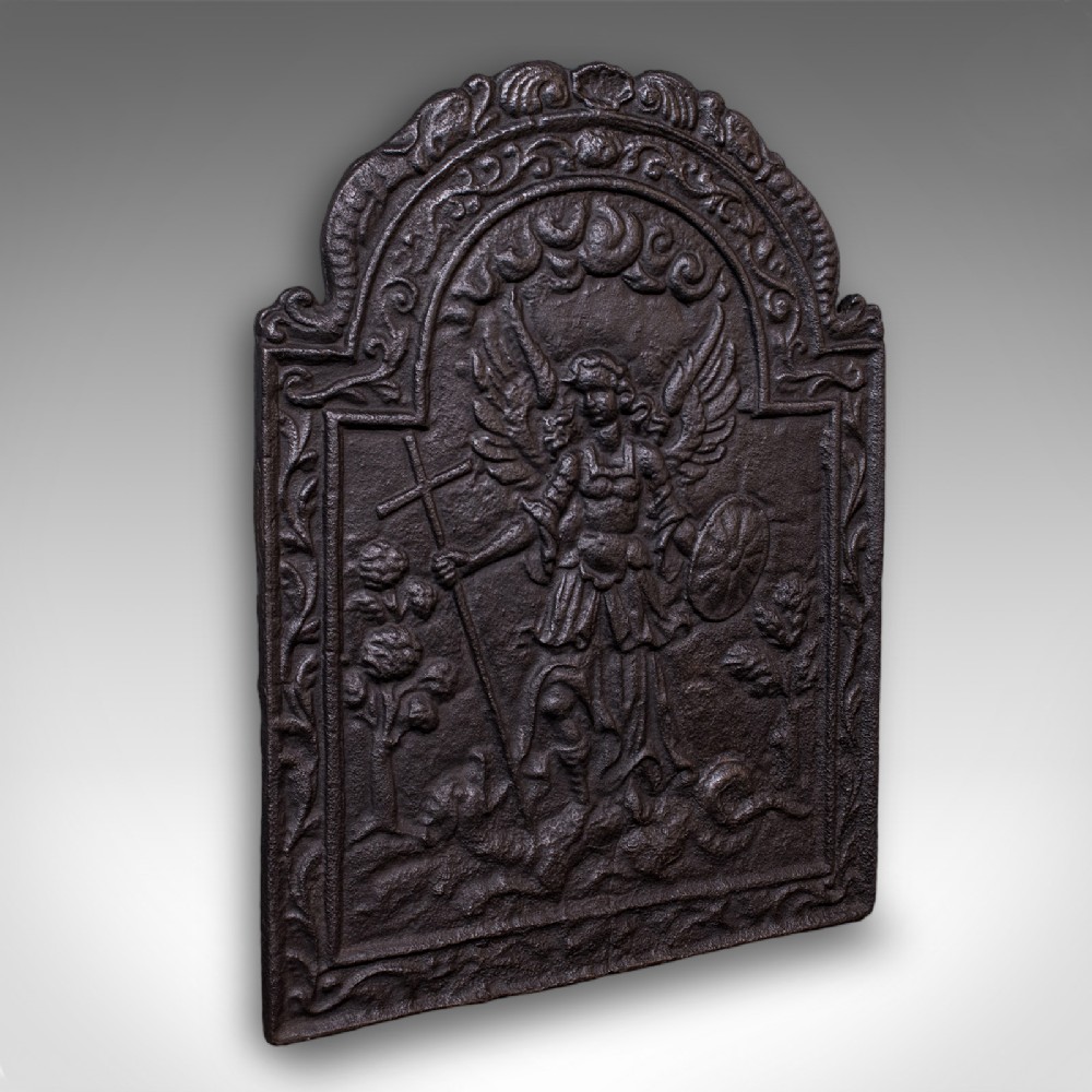 A heavy arched Victorian cast iron fireback featuring an embossed relief depicting an angelic figure with a cross and shield framed by detailed scrollwork borders.