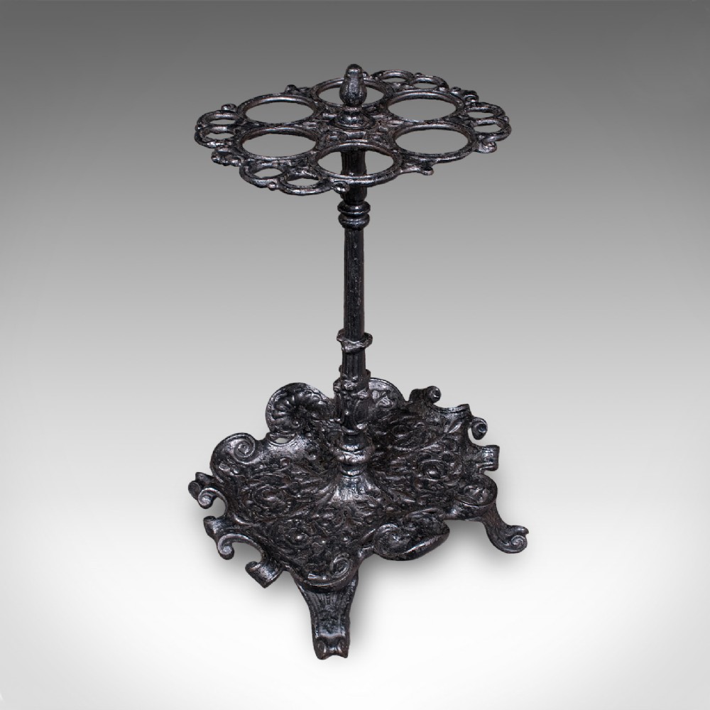 A Victorian cast iron stick stand featuring a central column, a six-loop upper ring, and a decorative scrollwork base with integrated drip tray in a dark finish.