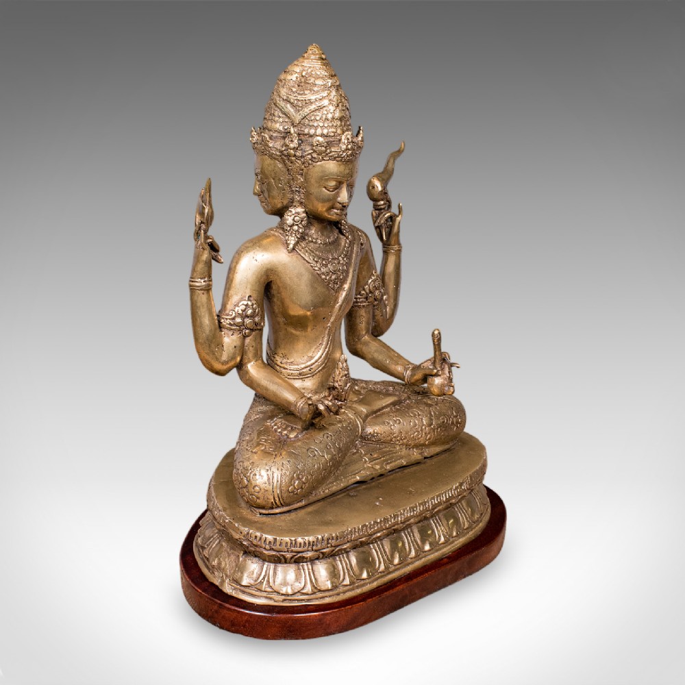 An antique bronze multi-headed and four-armed Balinese deity figure seated in lotus position on a lotus base, mounted on an oval wooden plinth.