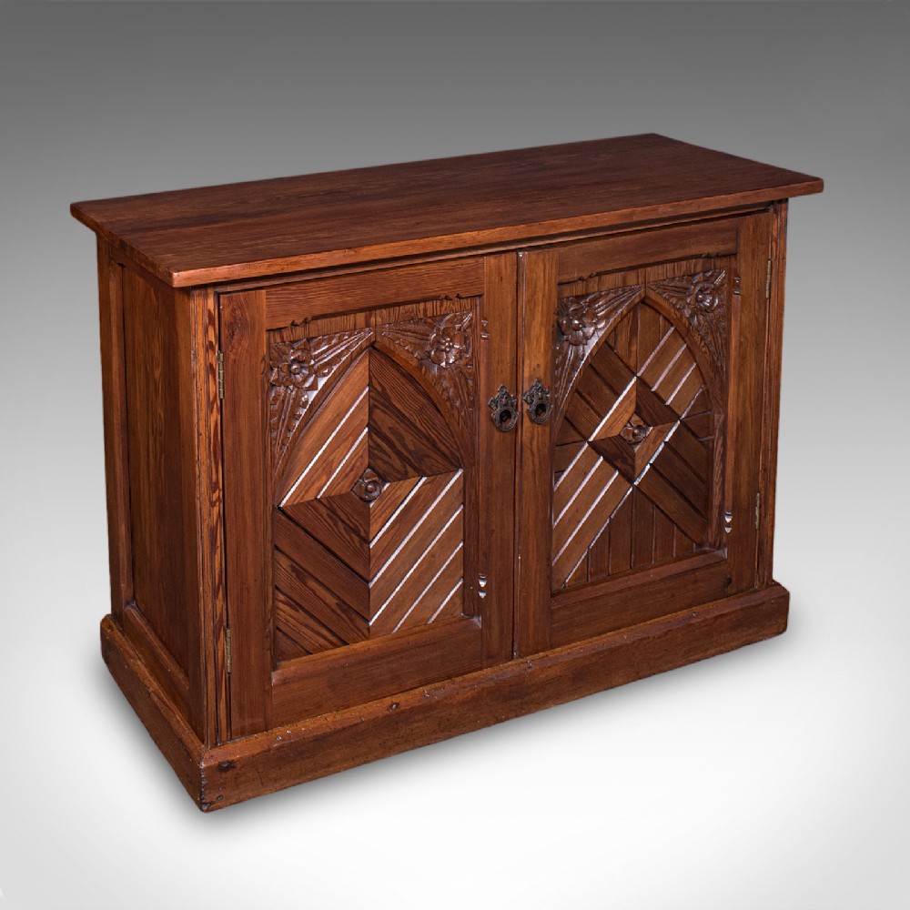 A low Victorian Gothic Revival wooden cabinet with double doors featuring carved pointed arches, geometric panelling, and twin metal drop handles.