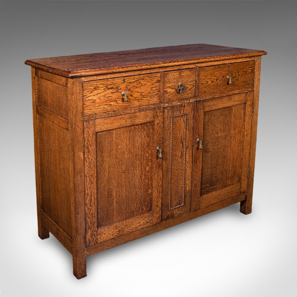 An antique Edwardian medium brown oak sideboard featuring three upper drawers with brass drop handles above panelled cupboard doors.