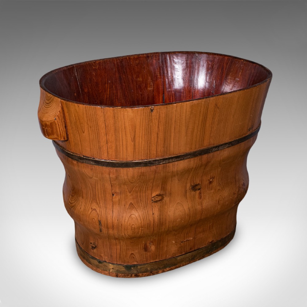 An oval late Victorian Dutch cedar waste paper basket with brass binding straps and carved side handles on a light background.