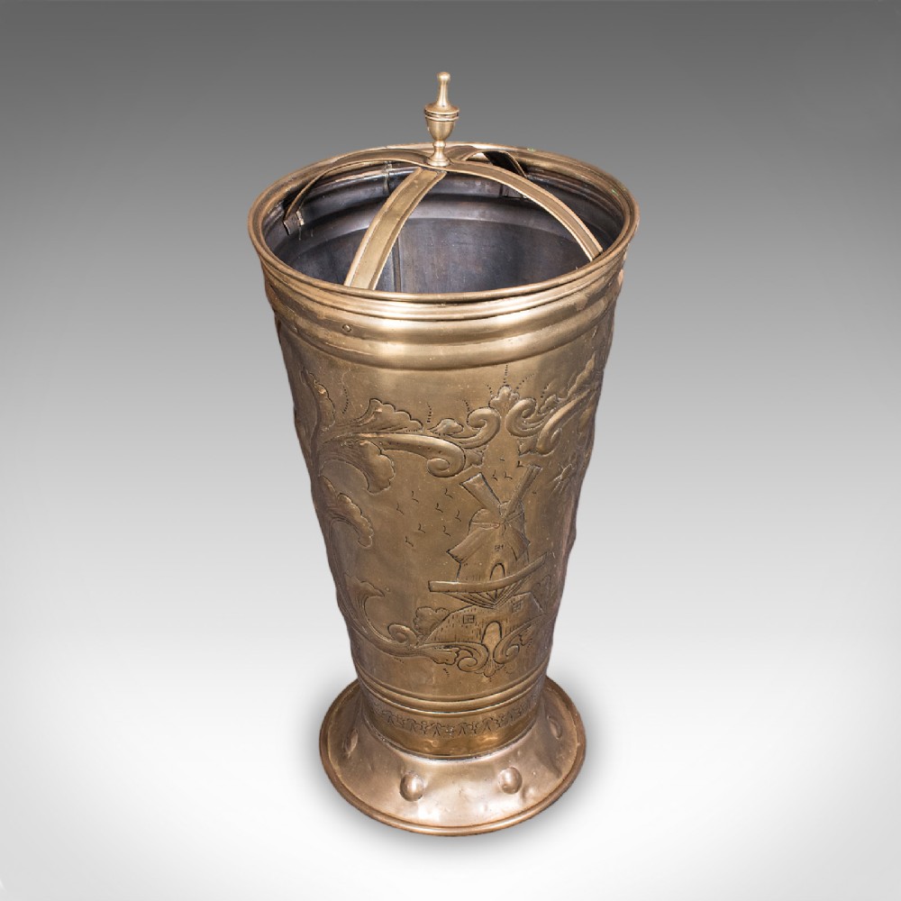 An antique tapered brass umbrella stand featuring embossed Dutch windmill motifs, top dividers with a turned brass finial, and a flared base.