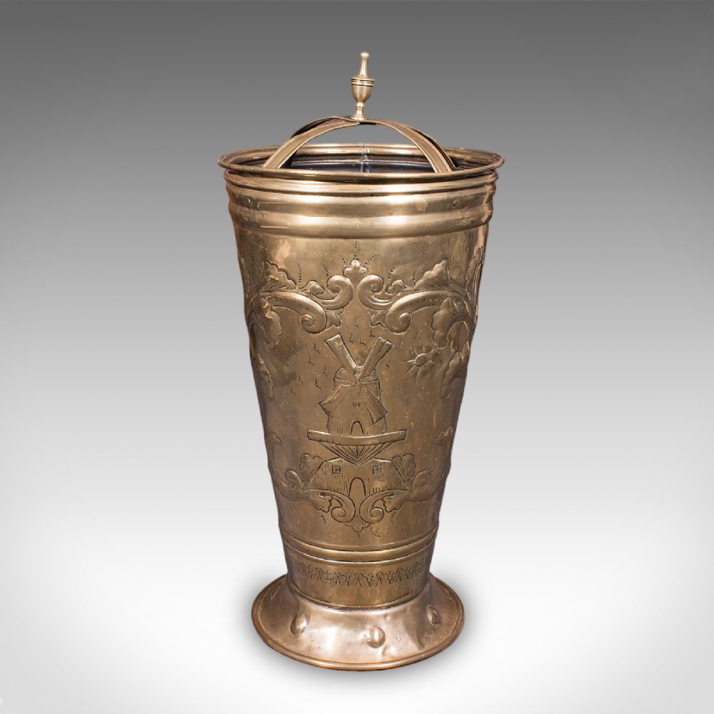 An antique tapered brass umbrella stand featuring embossed Dutch windmill motifs, top dividers with a turned brass finial, and a flared base. View II