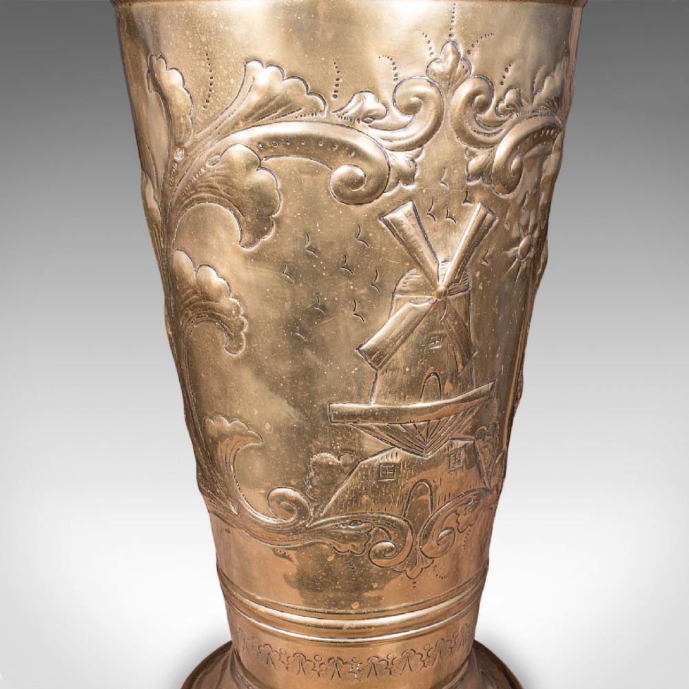 An antique tapered brass umbrella stand featuring embossed Dutch windmill motifs, top dividers with a turned brass finial, and a flared base. View VIII
