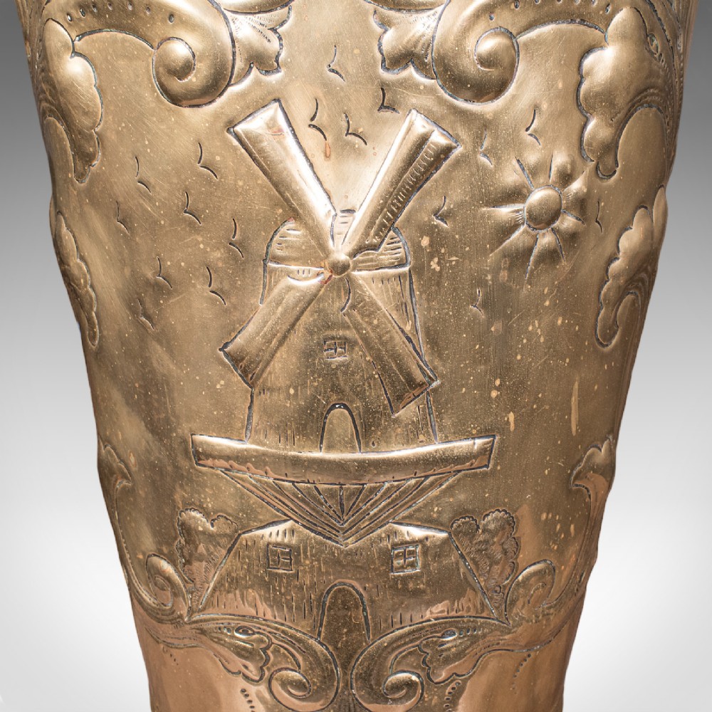 An antique tapered brass umbrella stand featuring embossed Dutch windmill motifs, top dividers with a turned brass finial, and a flared base. View IX