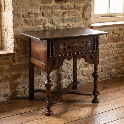 fig 1. A dark brown oak side table featuring a single carved drawer with brass handles, a decorative apron, turned baluster legs, and a shaped cross stretcher.