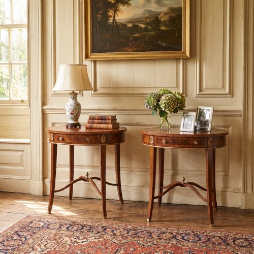 fig 1. A pair of oval flame mahogany side tables featuring brass rosette mounts, single drawers, curved legs, and an X-shaped stretcher with a brass finial.