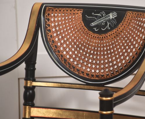 fig 2. A Regency style ebonised and parcel-gilt open armchair featuring a curved cane backrest with painted medallion detail, sweeping arms, and a patterned gold upholstered cushion.