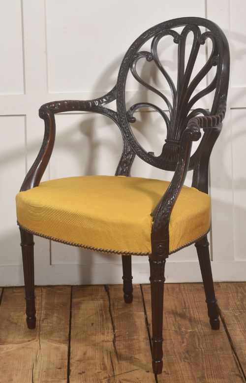 fig 1. A carved mahogany open armchair featuring an oval wheat-sheaf backrest, swept arms, and a yellow upholstered seat secured with brass studs.