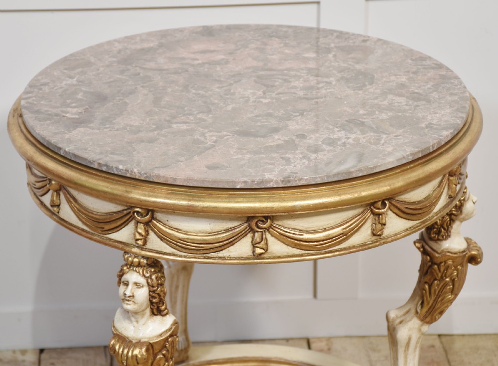A circular Neoclassical occasional table with a polished pink marble top, cream-painted frame, gold leaf drapery swags, and carved term legs ending in paw feet. View V