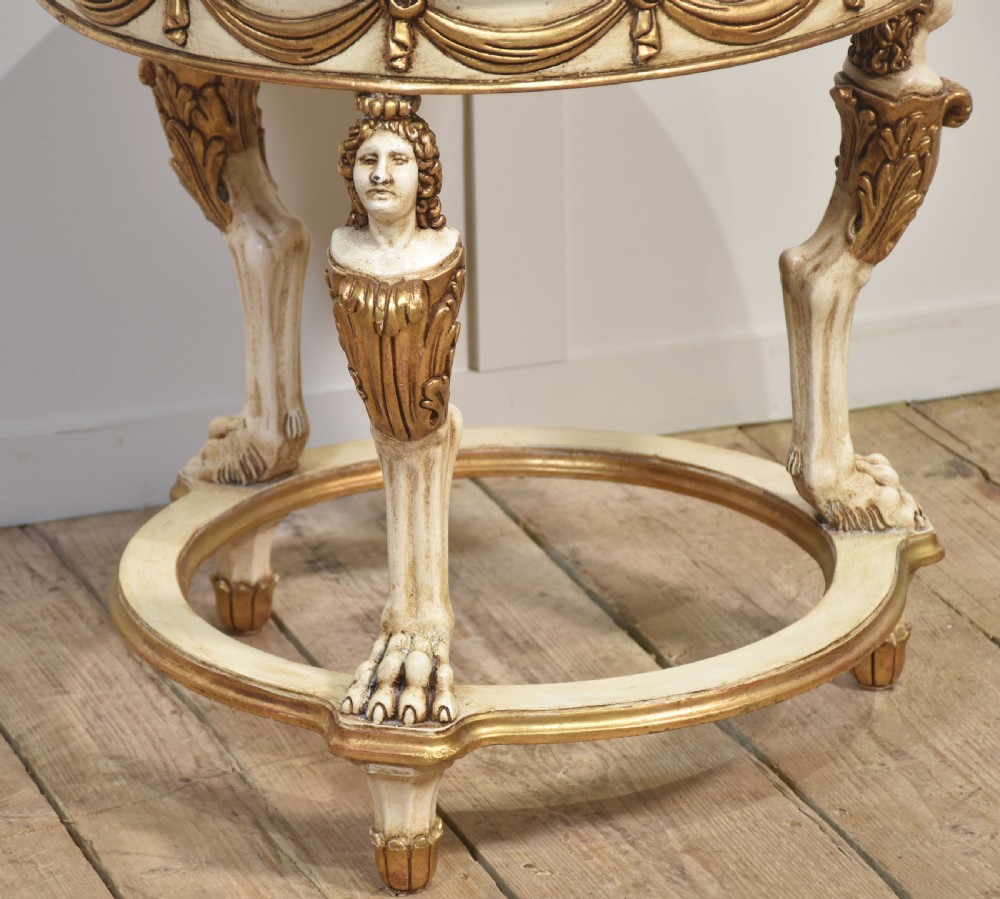 A circular Neoclassical occasional table with a polished pink marble top, cream-painted frame, gold leaf drapery swags, and carved term legs ending in paw feet. View VIII