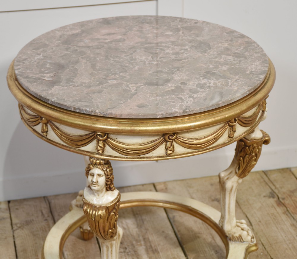 A circular Neoclassical occasional table with a polished pink marble top, cream-painted frame, gold leaf drapery swags, and carved term legs ending in paw feet. View IX