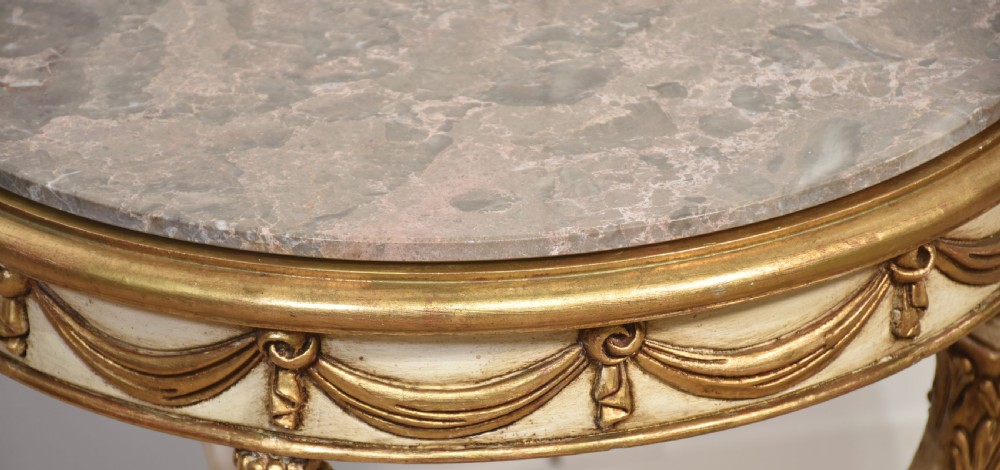 A circular Neoclassical occasional table with a polished pink marble top, cream-painted frame, gold leaf drapery swags, and carved term legs ending in paw feet. View X