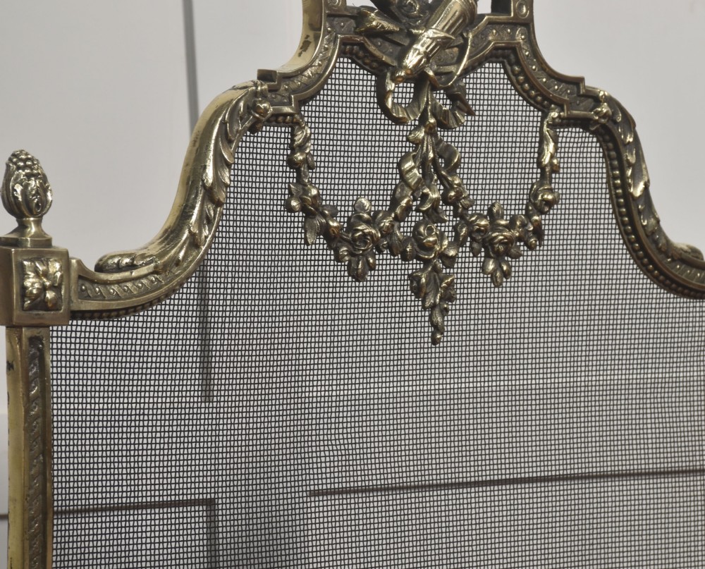 An ornate late 19th-century French gilt brass fire screen featuring a wire mesh panel decorated with a central urn motif, floral swags, and a top crest with a quiver and torch. View VII