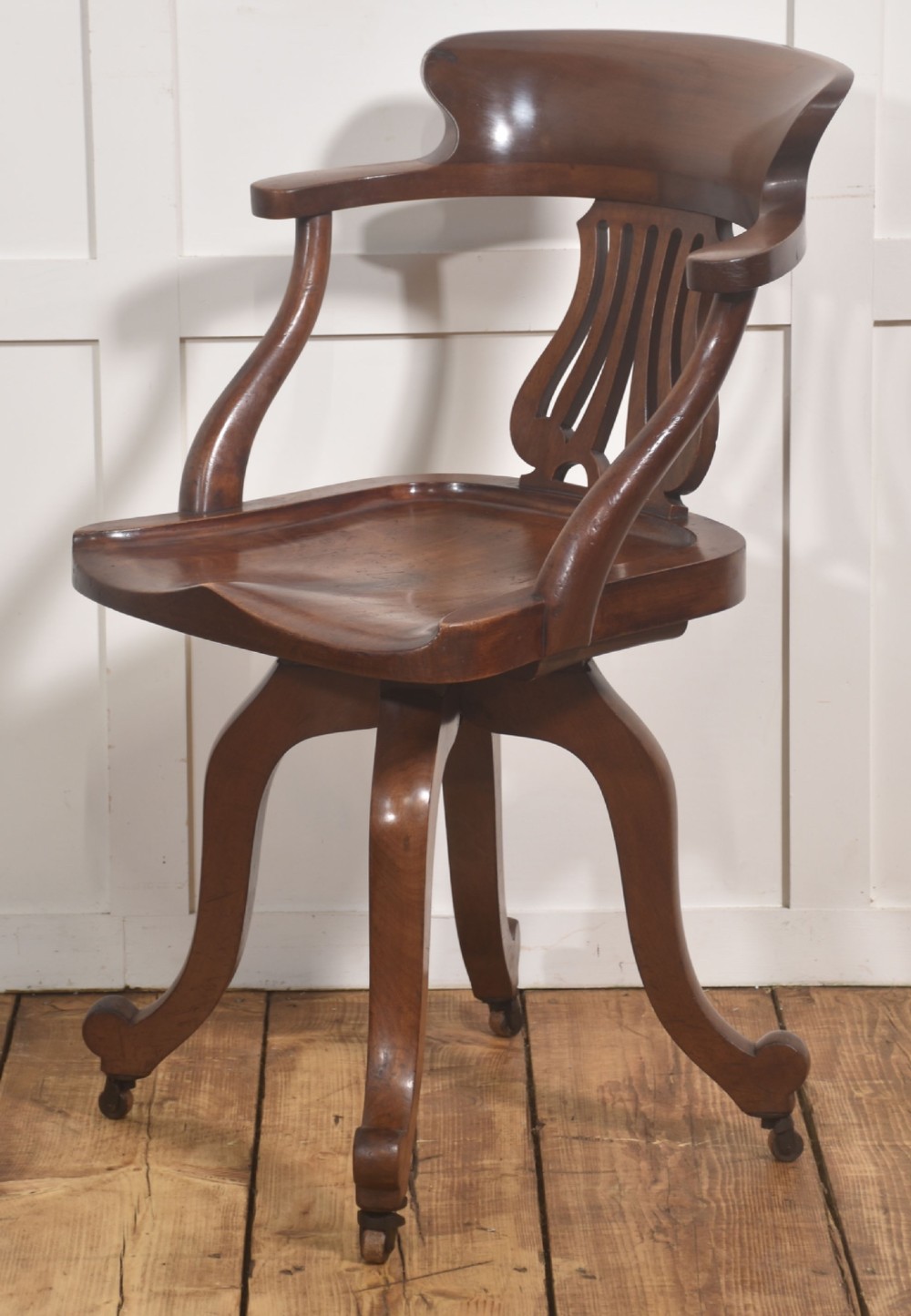 An antique mahogany revolving desk chair featuring a curved backrest, carved splat, shaped seat, and four splayed legs fitted with castors. View V