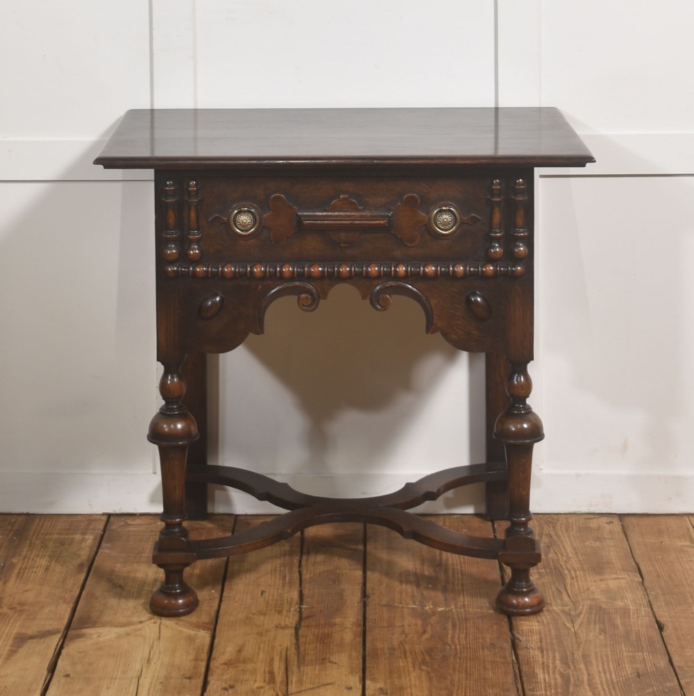 A dark brown oak side table featuring a single carved drawer with brass handles, a decorative apron, turned baluster legs, and a shaped cross stretcher.
