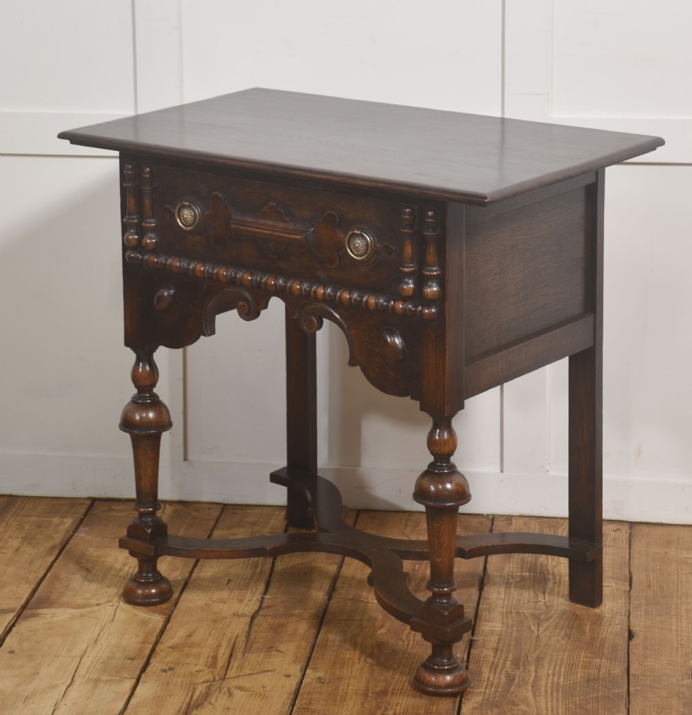 A dark brown oak side table featuring a single carved drawer with brass handles, a decorative apron, turned baluster legs, and a shaped cross stretcher. View V