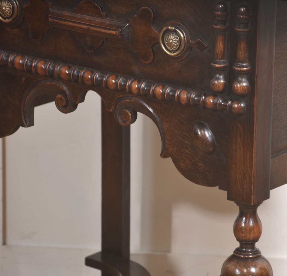 A dark brown oak side table featuring a single carved drawer with brass handles, a decorative apron, turned baluster legs, and a shaped cross stretcher. View VI