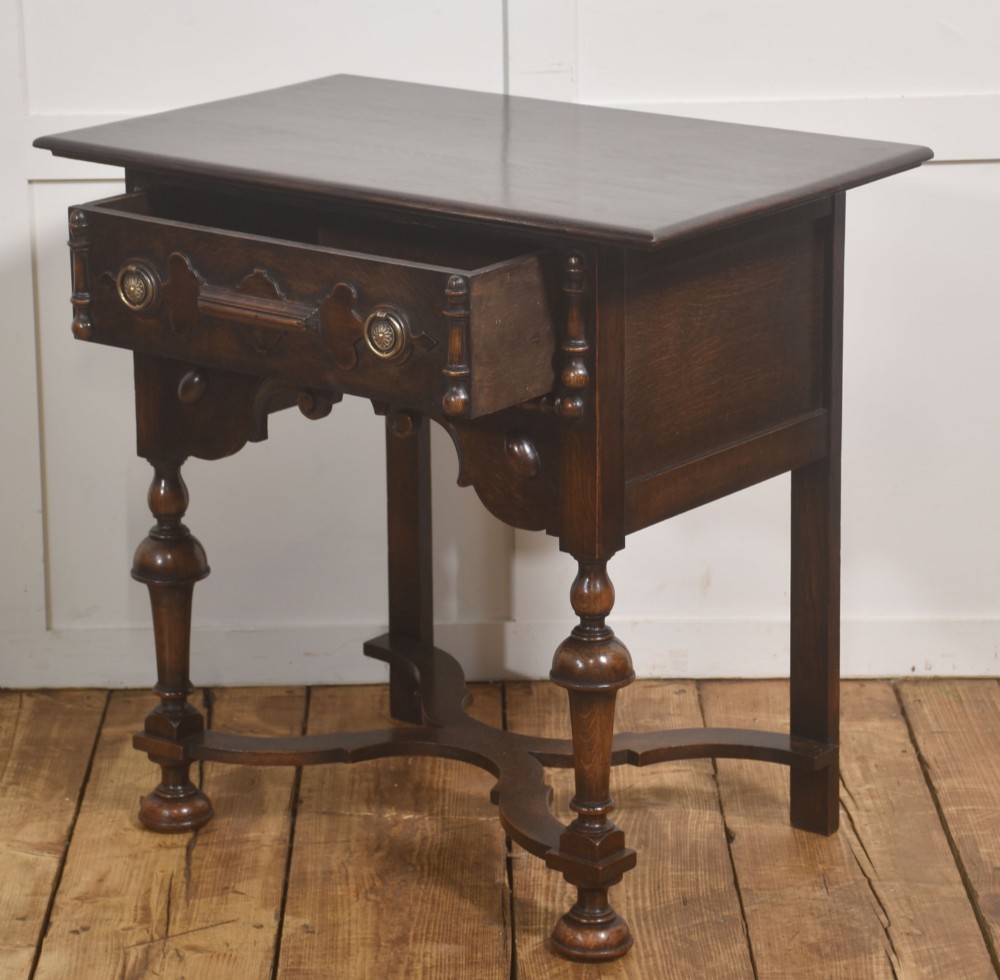 A dark brown oak side table featuring a single carved drawer with brass handles, a decorative apron, turned baluster legs, and a shaped cross stretcher. View IX