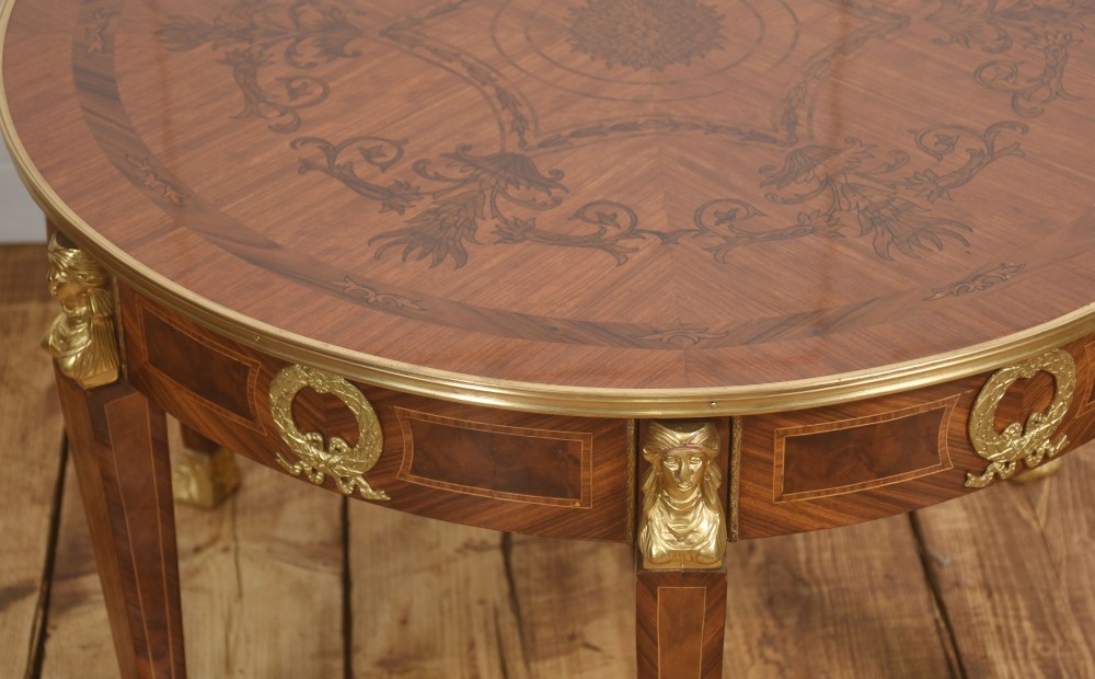 A round Louis XVI style coffee table featuring wood marquetry, warm brown veneers, tapered square legs, and decorative gilt brass female bust mounts. View VI