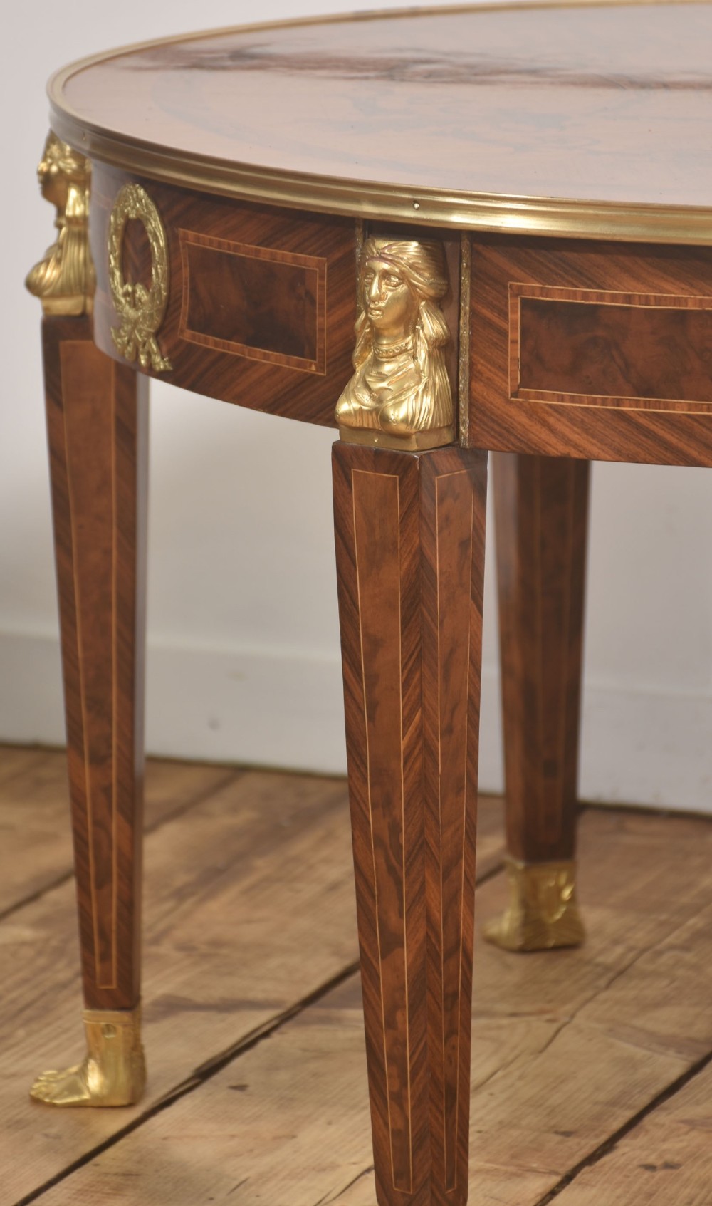 A round Louis XVI style coffee table featuring wood marquetry, warm brown veneers, tapered square legs, and decorative gilt brass female bust mounts. View X