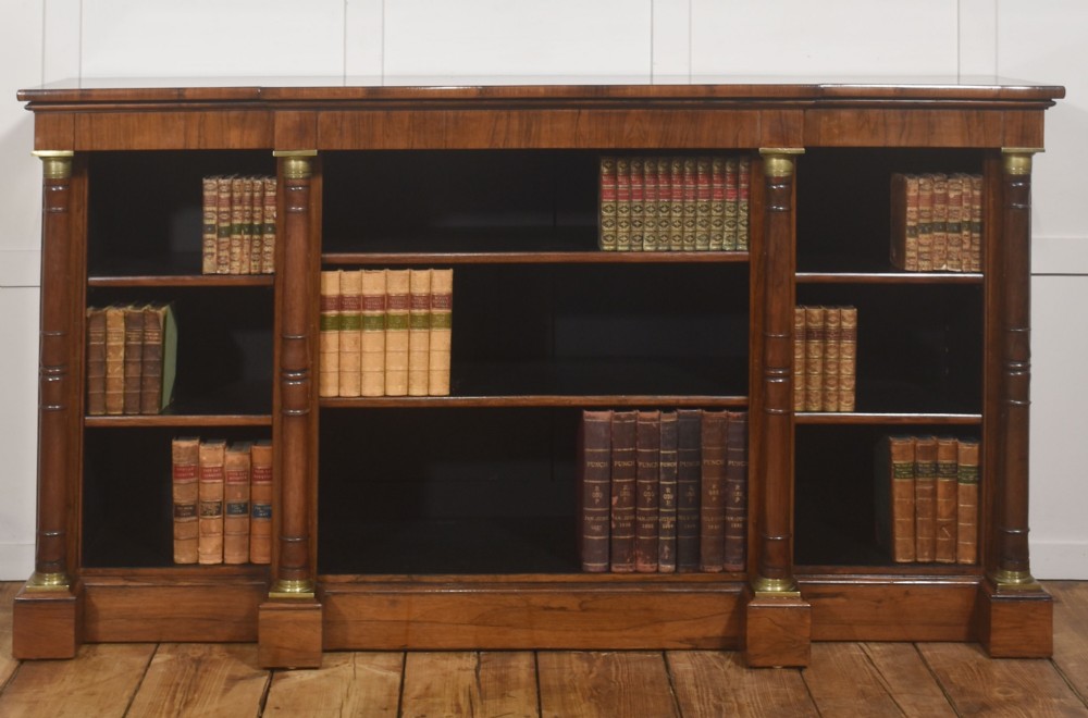 A Regency period open breakfront bookcase crafted from dark rosewood with brass mounted column pilasters and adjustable shelves.