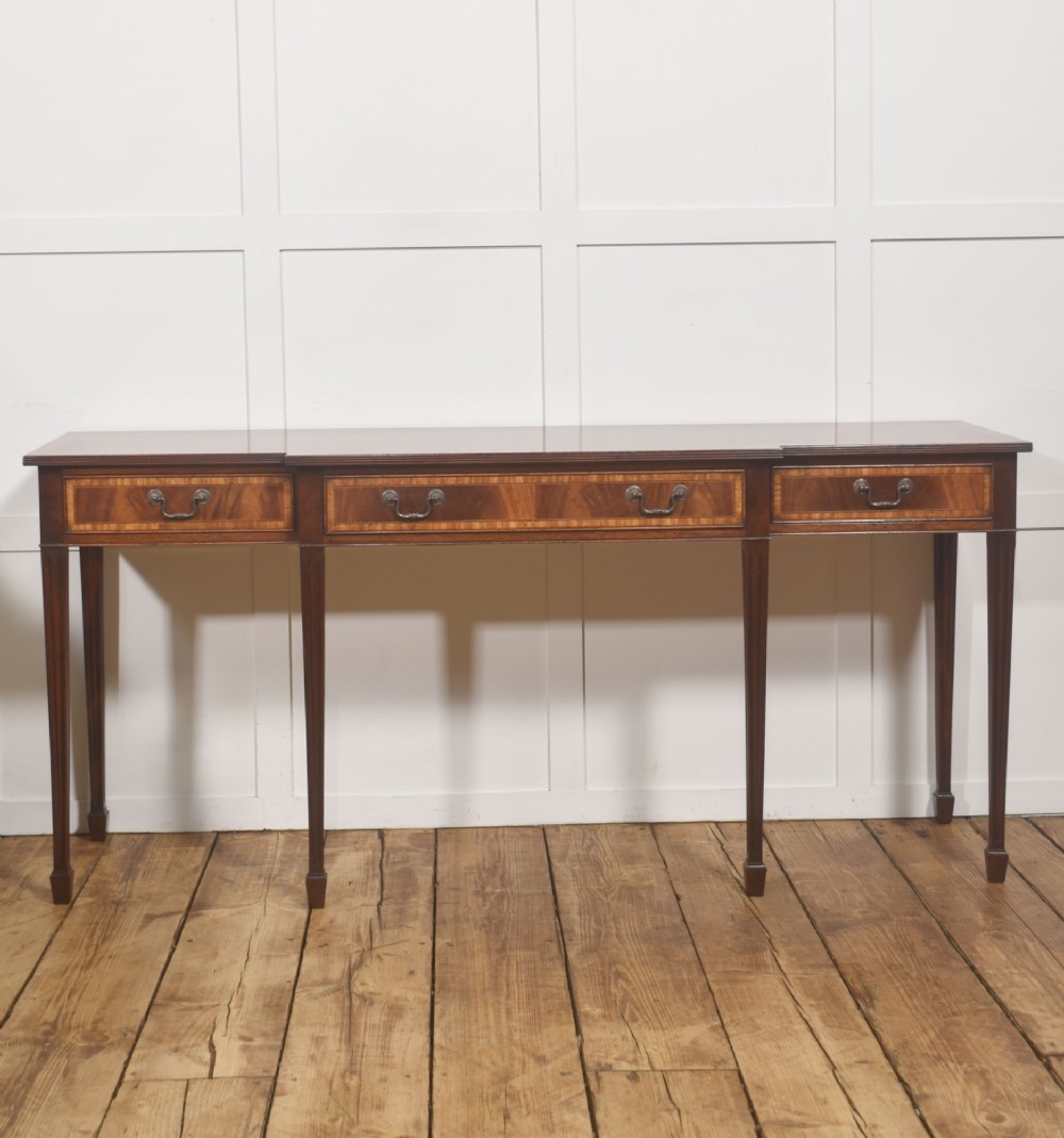 A mahogany breakfront hall table featuring satinwood crossbanding, three drawers with brass handles, and four tapered square legs.