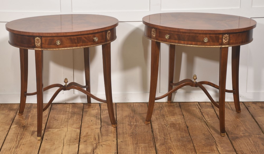 A pair of oval flame mahogany side tables featuring brass rosette mounts, single drawers, curved legs, and an X-shaped stretcher with a brass finial. View V